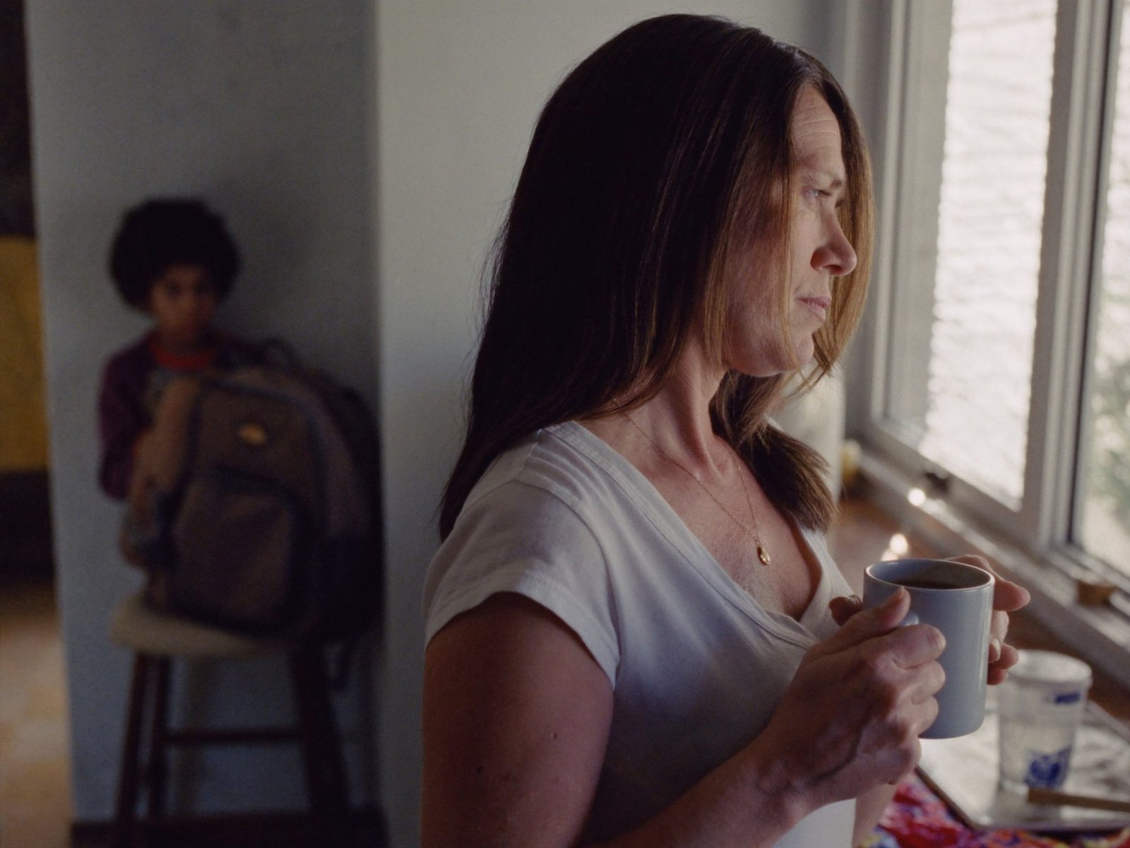 A mother at a kitchen window with a cup of coffee, deep in thought, a child behind her