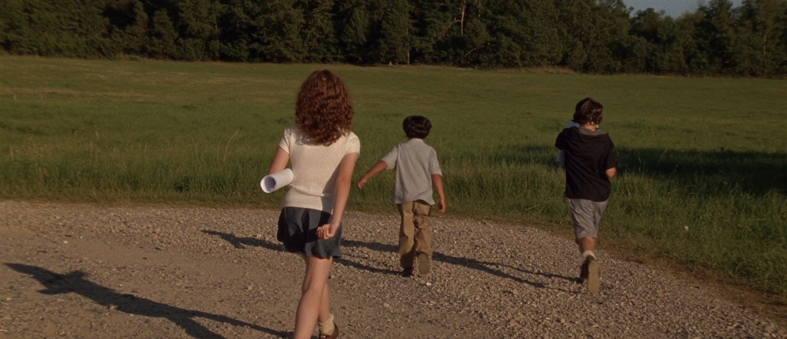 Three children walking toward an open field at golden hour, one carrying a rolled blueprint
